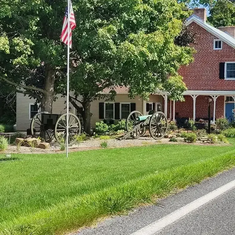 Historic red brick house with white trim, American flag, and antique cannons behind a green lawn.