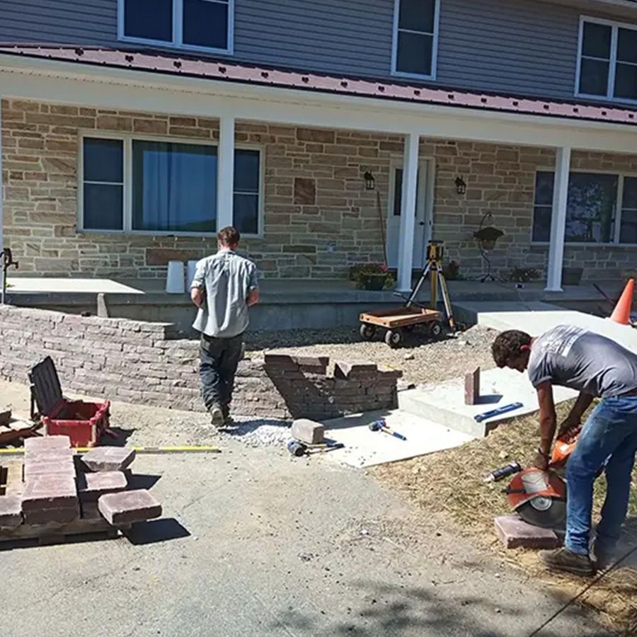 Workers laying pavers and leveling a stone patio beside a brick building