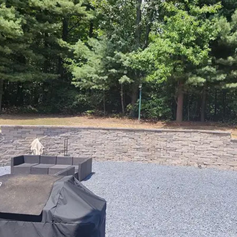 Stone patio with black fire pit, gravel ground, and wooded backyard behind a retaining wall