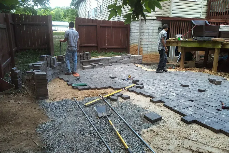 Workers laying gray pavers in a backyard patio with tools and stacked materials.