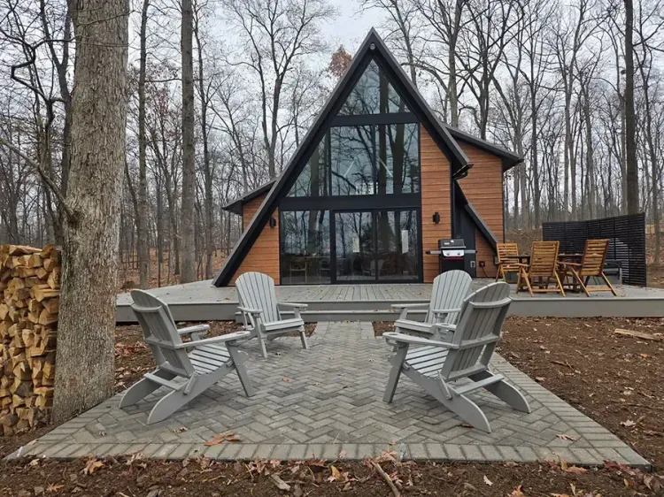 Modern A-frame cabin with large windows, surrounded by leafless trees and a paved patio with outdoor chairs.