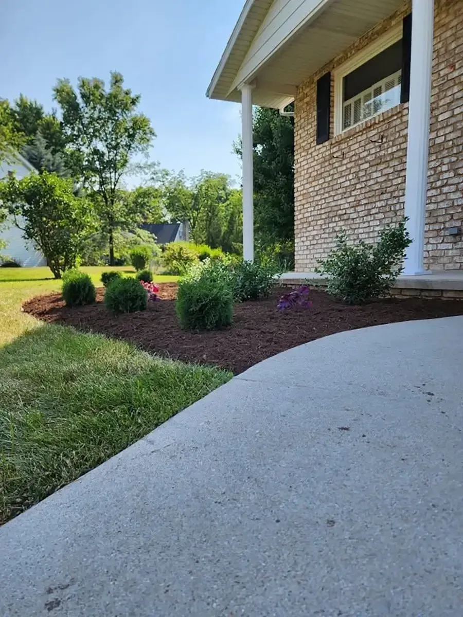 Curved concrete walkway beside a brick house with mulched flower beds, shrubs, and a green lawn