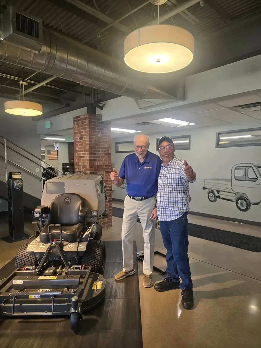 Two people posing beside a black riding mower in a showroom, one giving a thumbs-up.