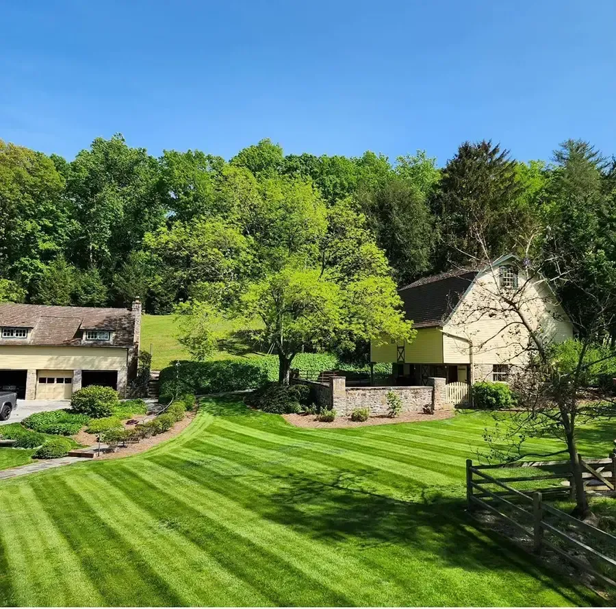 Sunny countryside house with striped green lawn, stone walls, and trees under a blue sky