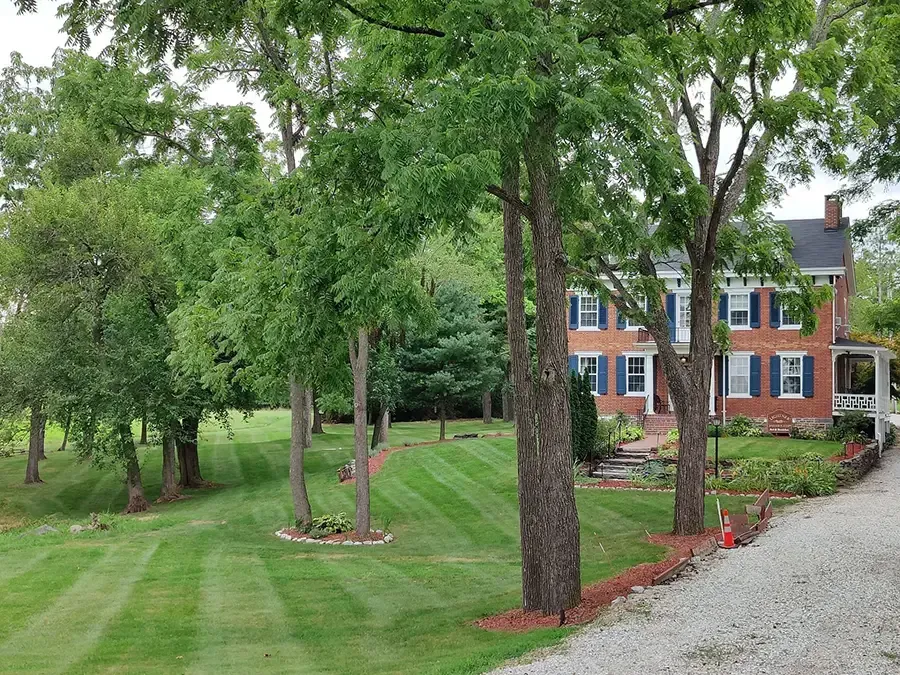 Tree-lined lawn beside a brick house with a gravel driveway and flower beds