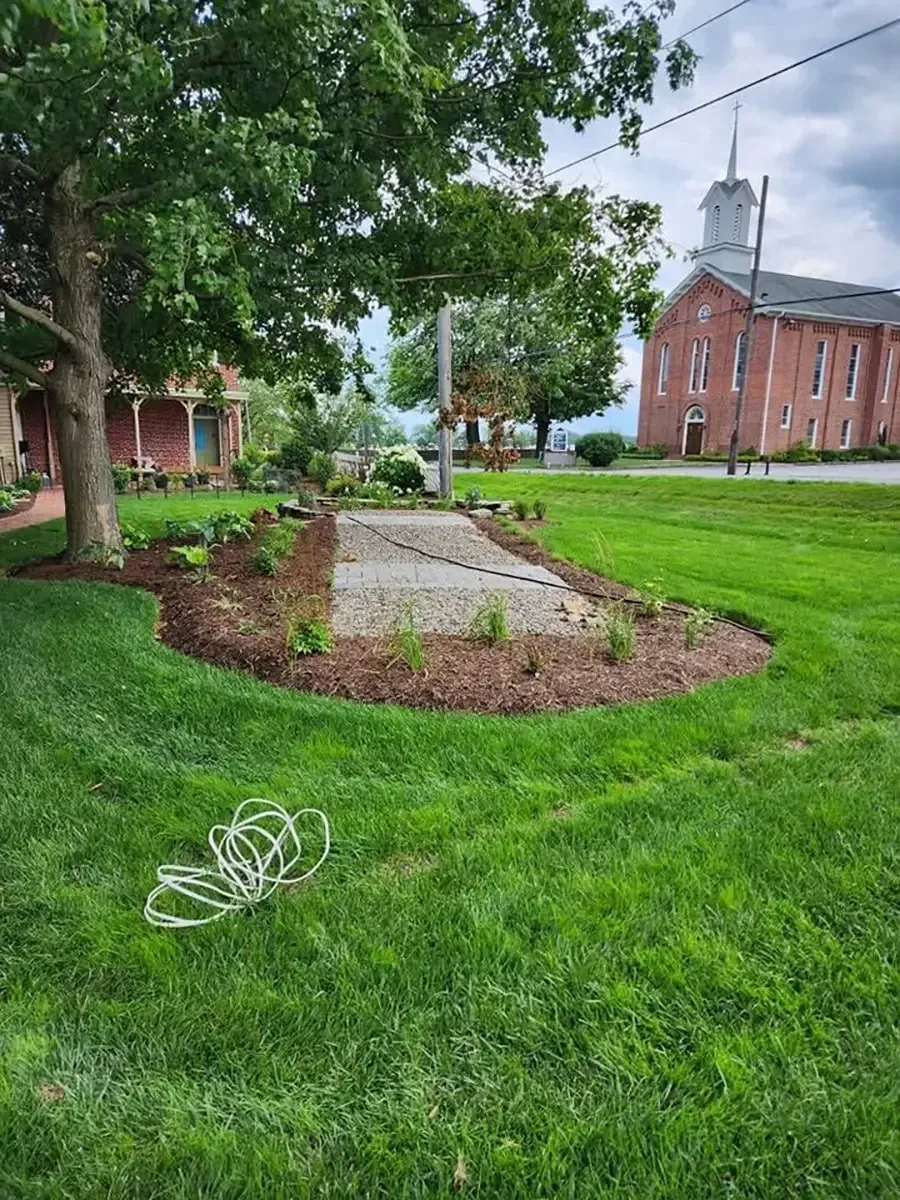A grassy campus lawn with a small garden bed, trees, and a brick building with a white tower in the background