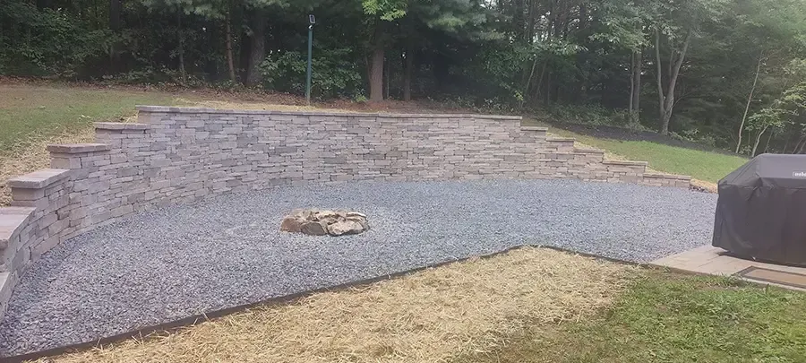 Gravel patio with a fire pit, bordered by greenery and a wooded backdrop.