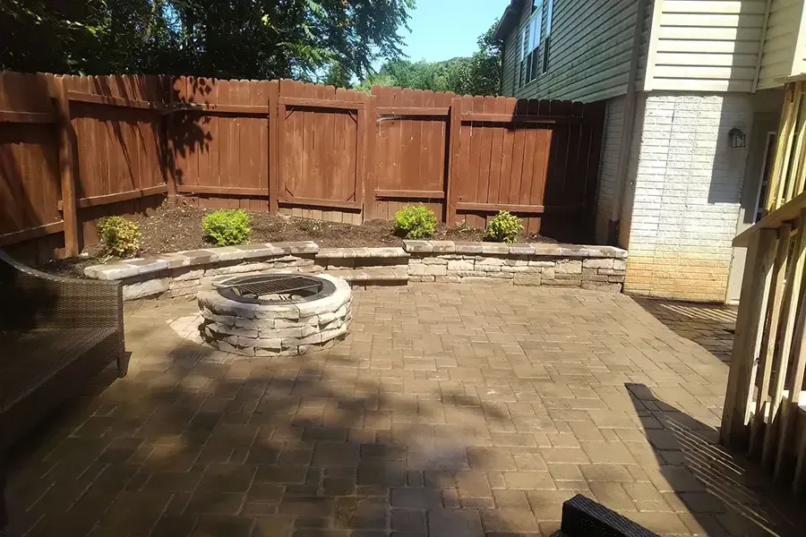 Brick patio with a circular fire pit, wooden fence, and potted plants in bright sunlight