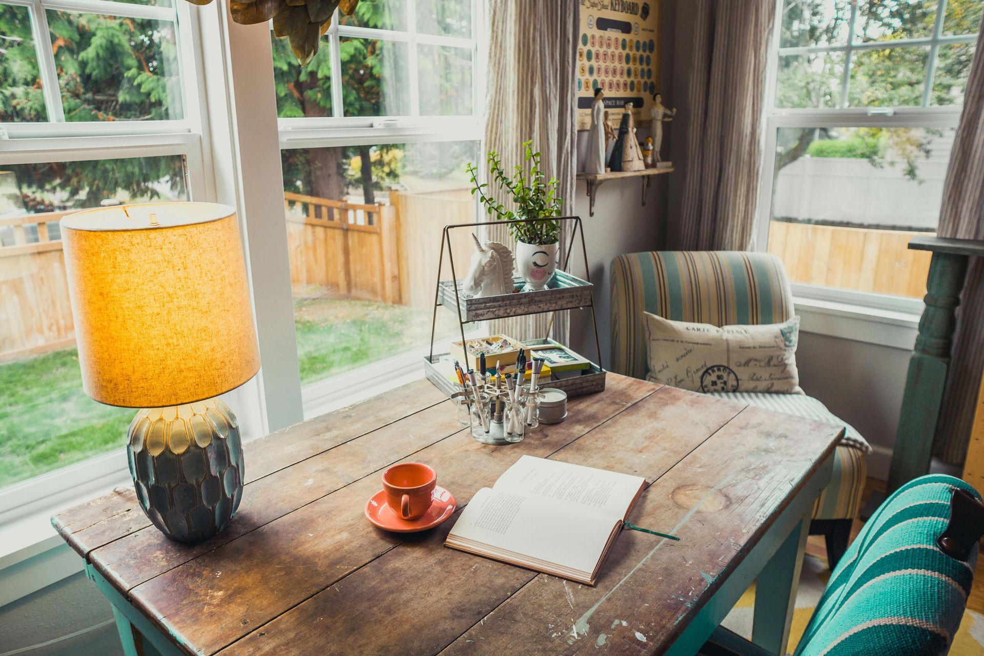 Cozy workspace by a window, wooden desk with lamp, chair, and open book. Plants and garden view.