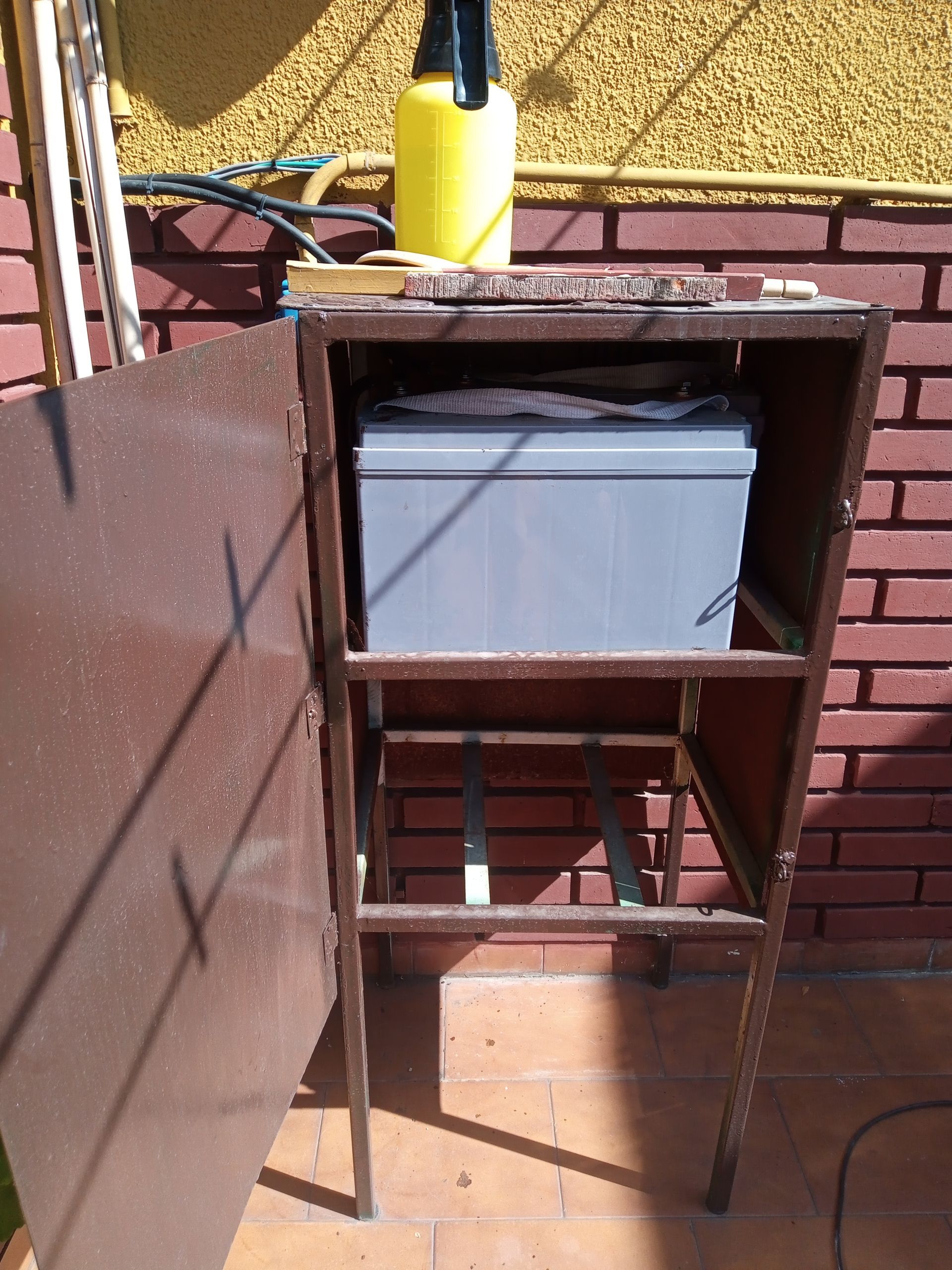 Un carrito de metal oxidado con una nevera portátil gris en un patio soleado, con una botella amarilla encima.