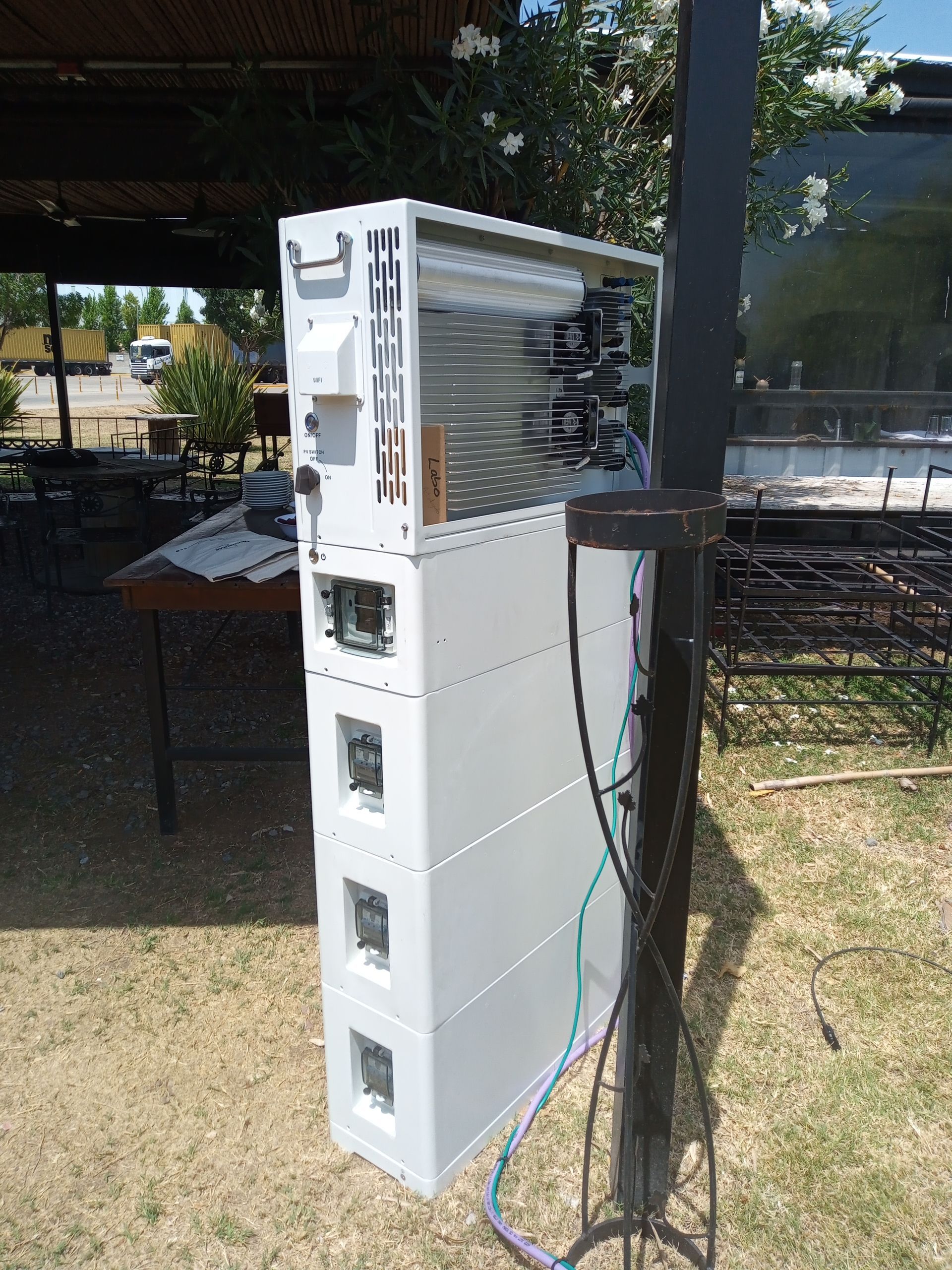 Pila blanca de electrodomésticos/aparatos electrónicos al aire libre, junto a un poste negro bajo un toldo.