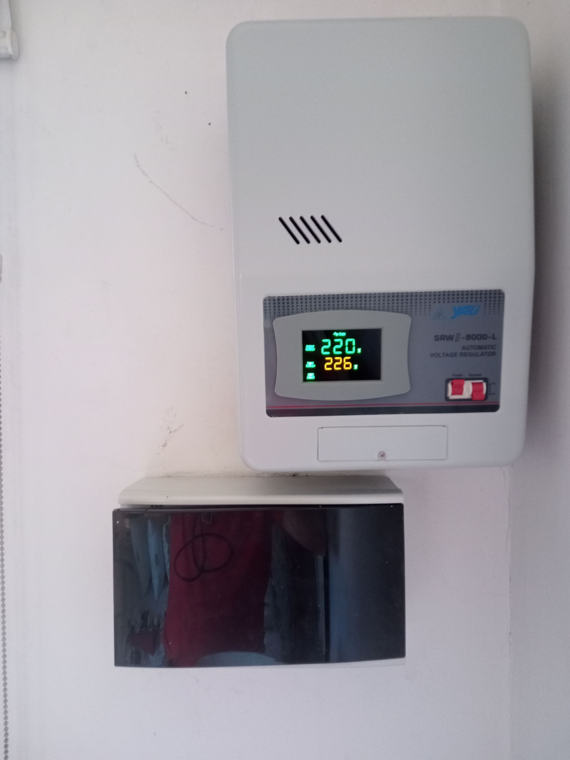 Termostato/unidad de control blanca de montaje en pared con pantalla digital, situada encima de una caja eléctrica negra.