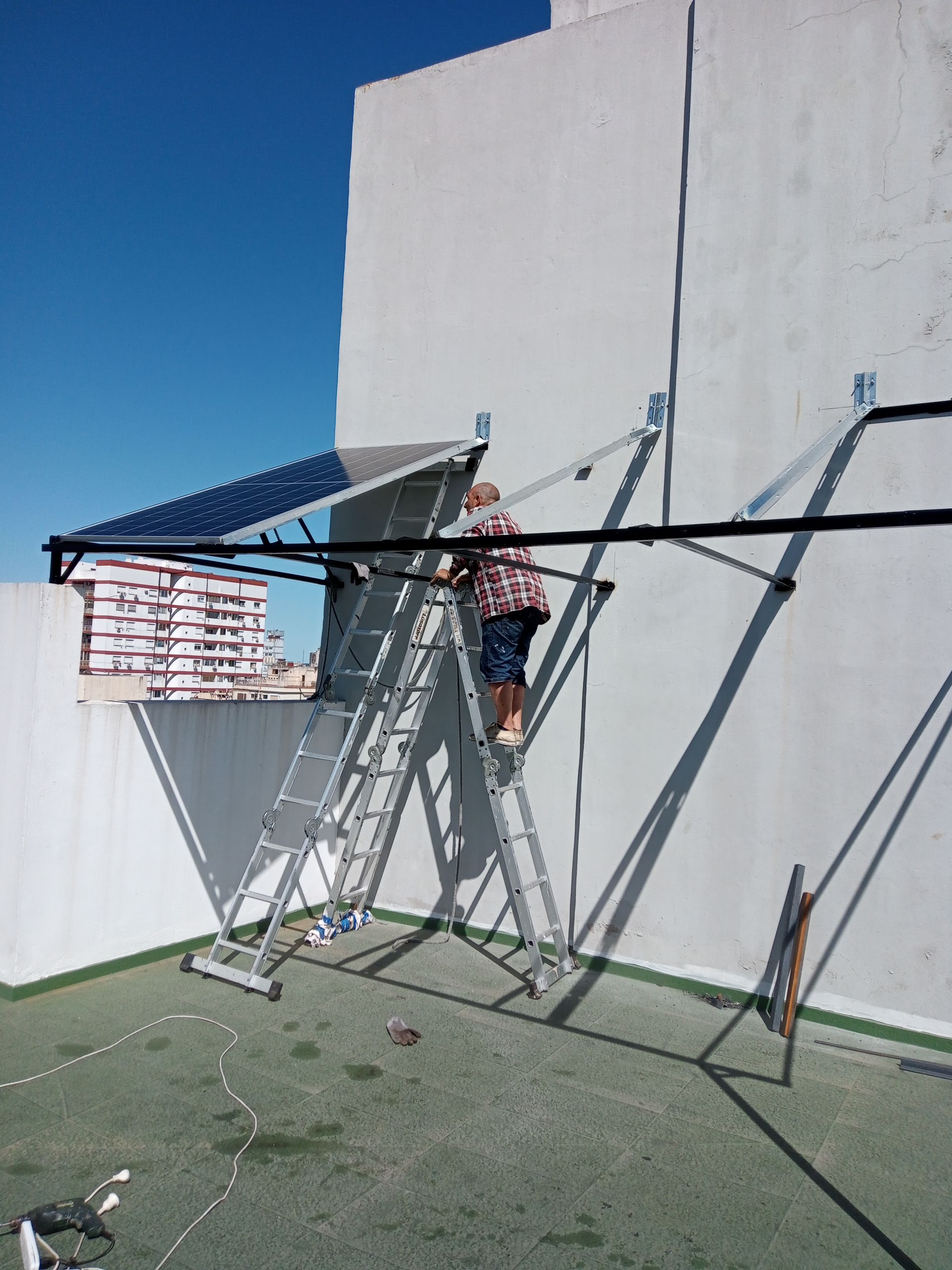 Un trabajador subido a una escalera ajusta un toldo negro contra una pared blanca en una azotea.