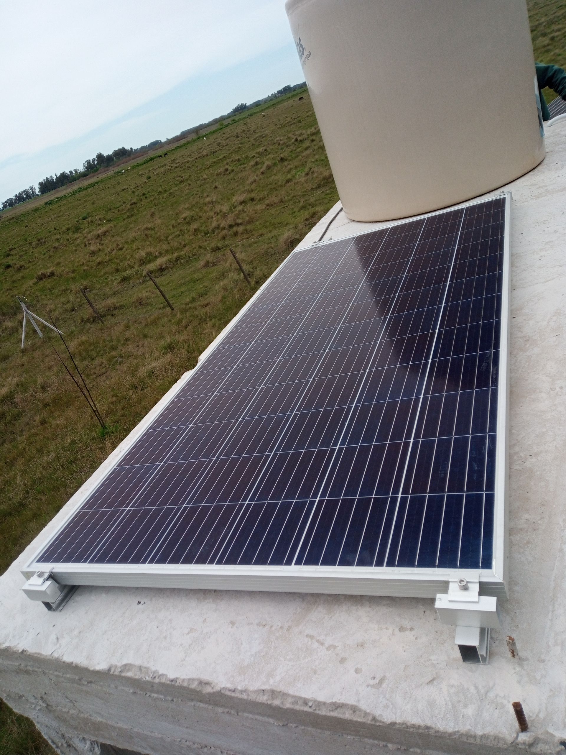 Panel solar instalado en un tejado junto a un depósito cilíndrico blanco, con vistas a un campo de hierba.