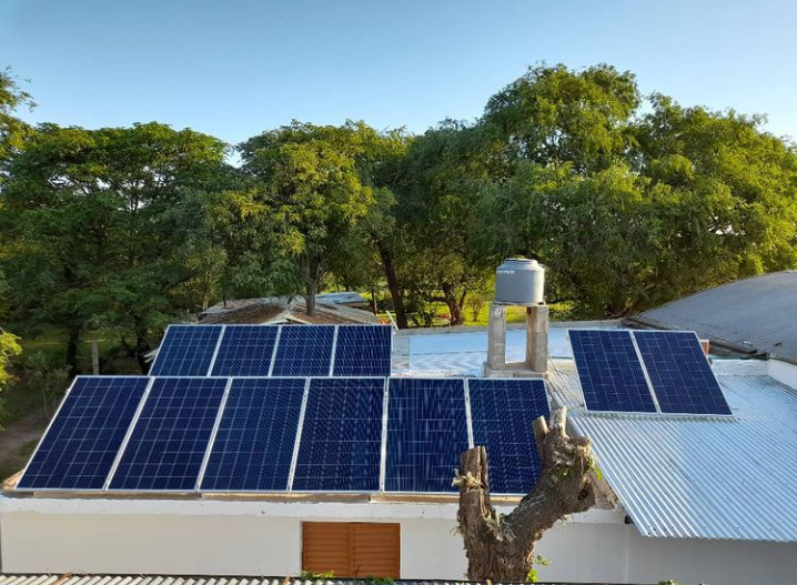 Paneles solares en el tejado de un edificio blanco, con árboles y cielo azul al fondo.