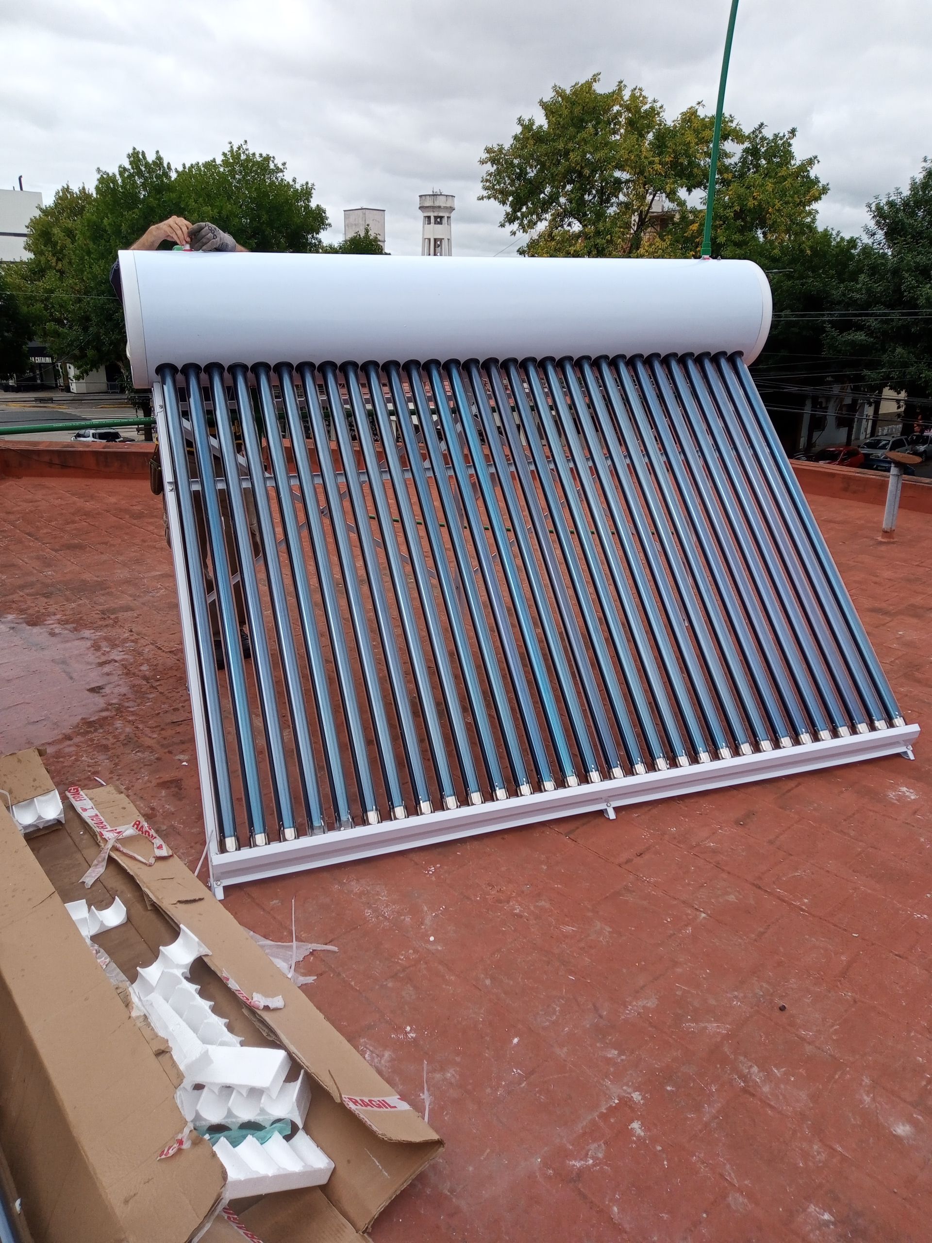 Colector solar térmico con tubos brillantes apoyado al aire libre sobre una superficie roja, junto a una caja abierta.