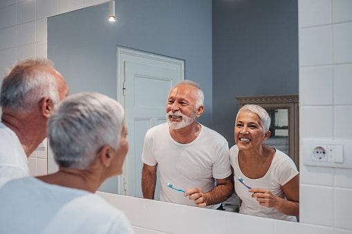 An elderly couple is brushing their teeth in front of a bathroom mirror.