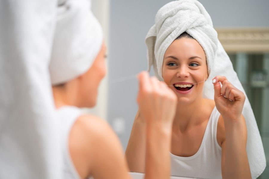 A woman with a towel wrapped around her head is flossing her teeth in front of a mirror.