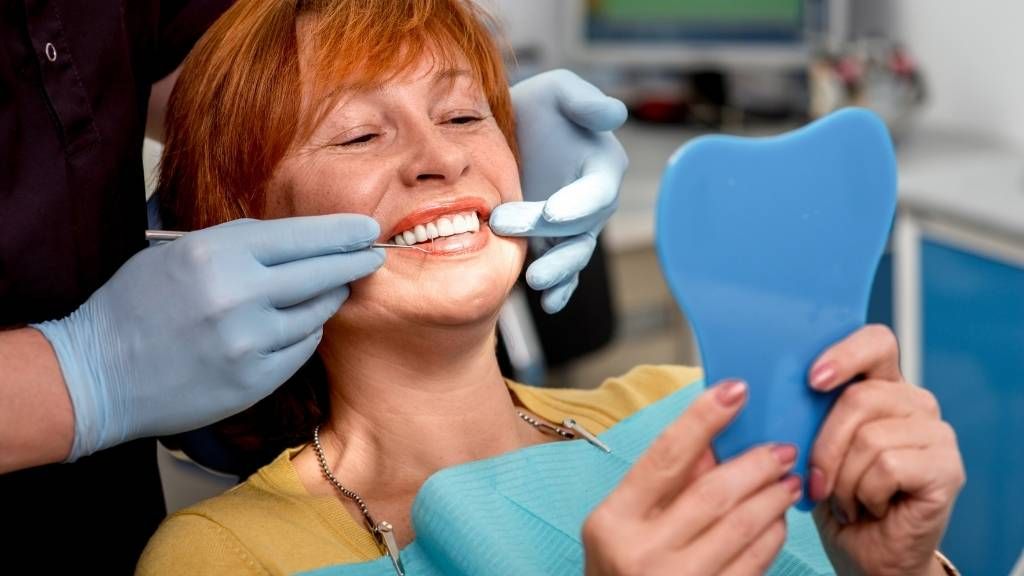 A woman is sitting in a dental chair looking at her teeth in a mirror.