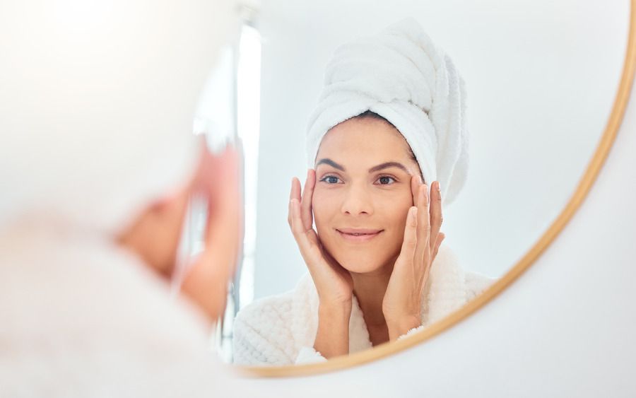 A woman with a towel wrapped around her head is looking at her face in a mirror.