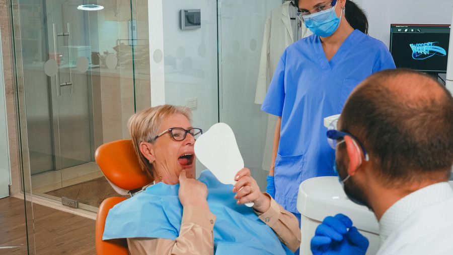 A woman is sitting in a dental chair looking at her teeth in a mirror.