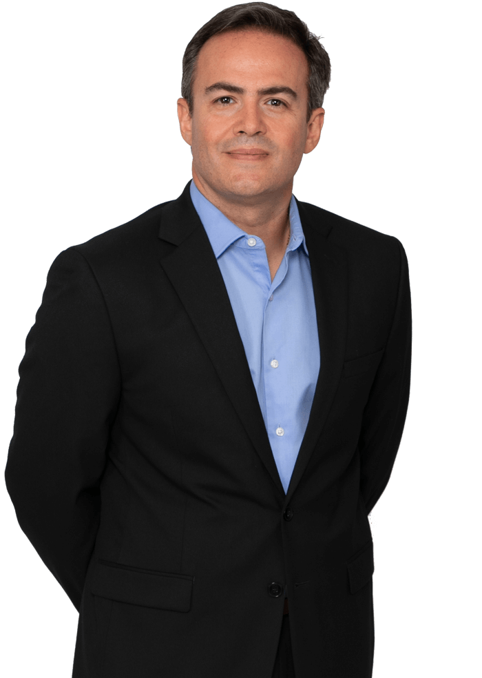 Man in a black suit and blue shirt, hands clasped behind his back, smiles slightly.