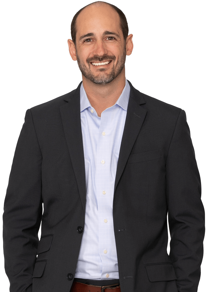 Man in a blue button-down shirt and dark blazer smiling, hands in pockets, against a white background.