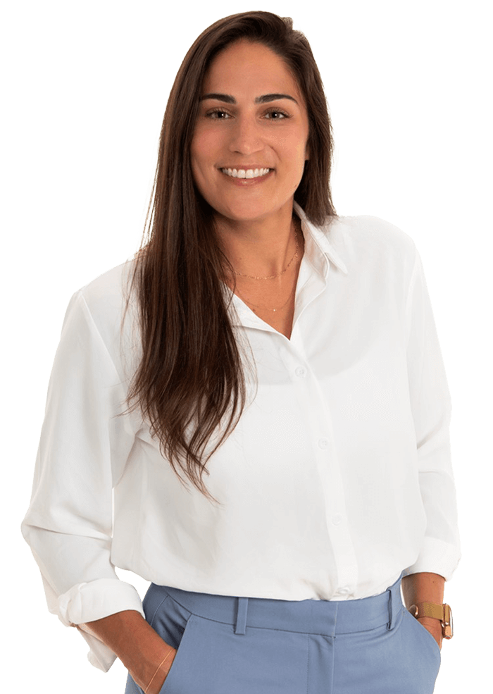 Woman with long brown hair, wearing a white button-down shirt and blue pants, smiling, hands in pockets.