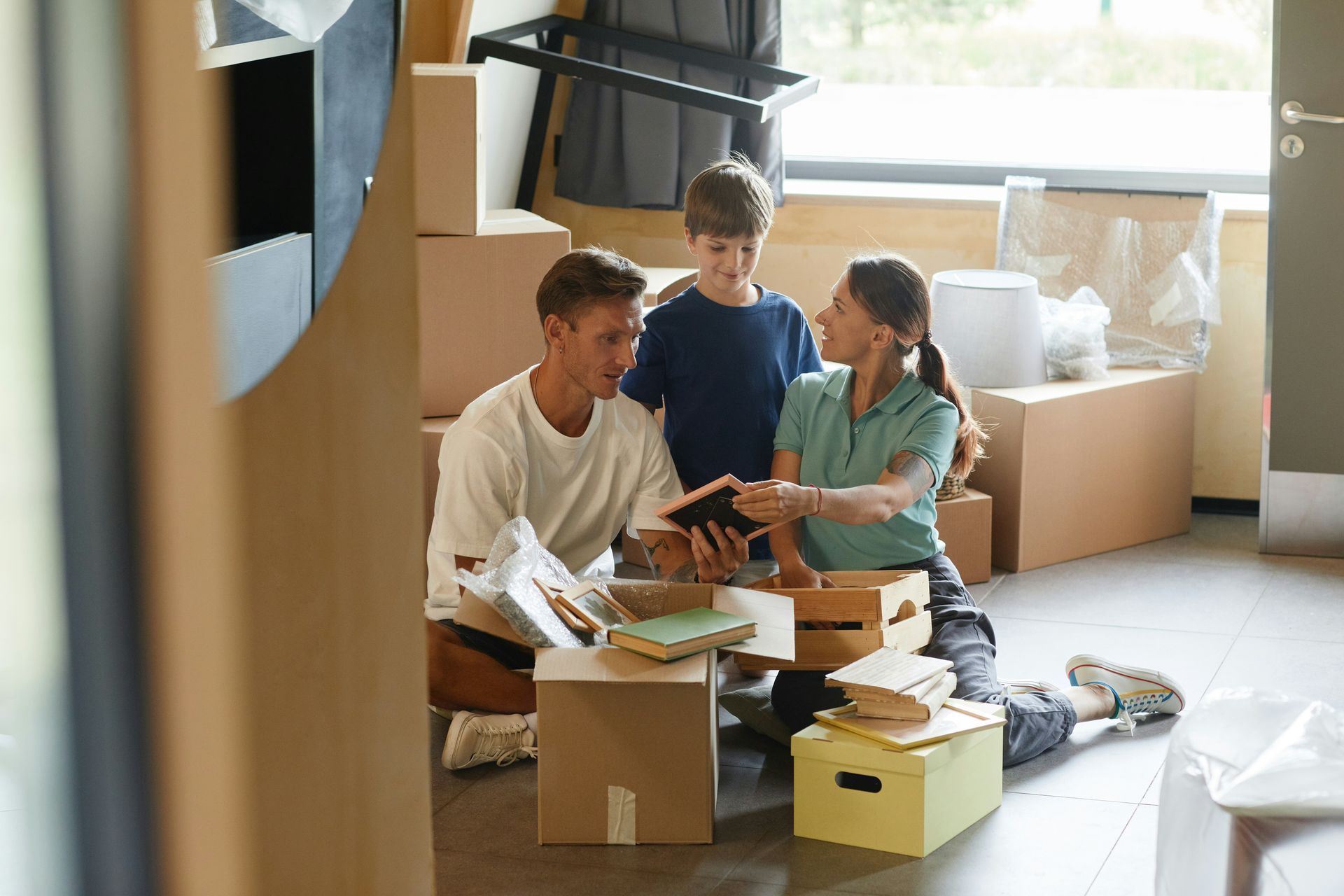 Family unpacking boxes in a new home; boy smiles, parents sort items; cardboard boxes surround.