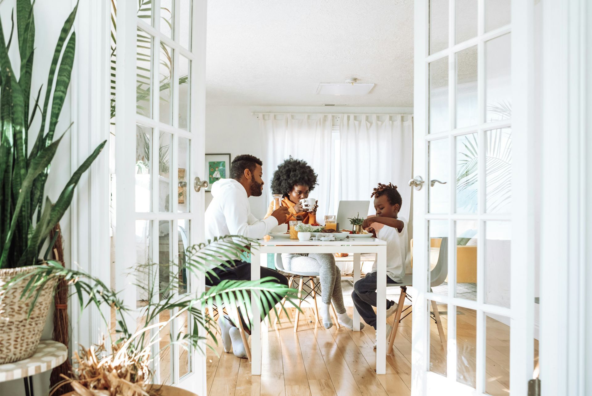 Family sitting at a white table using a laptop, in a bright room with French doors and plants.