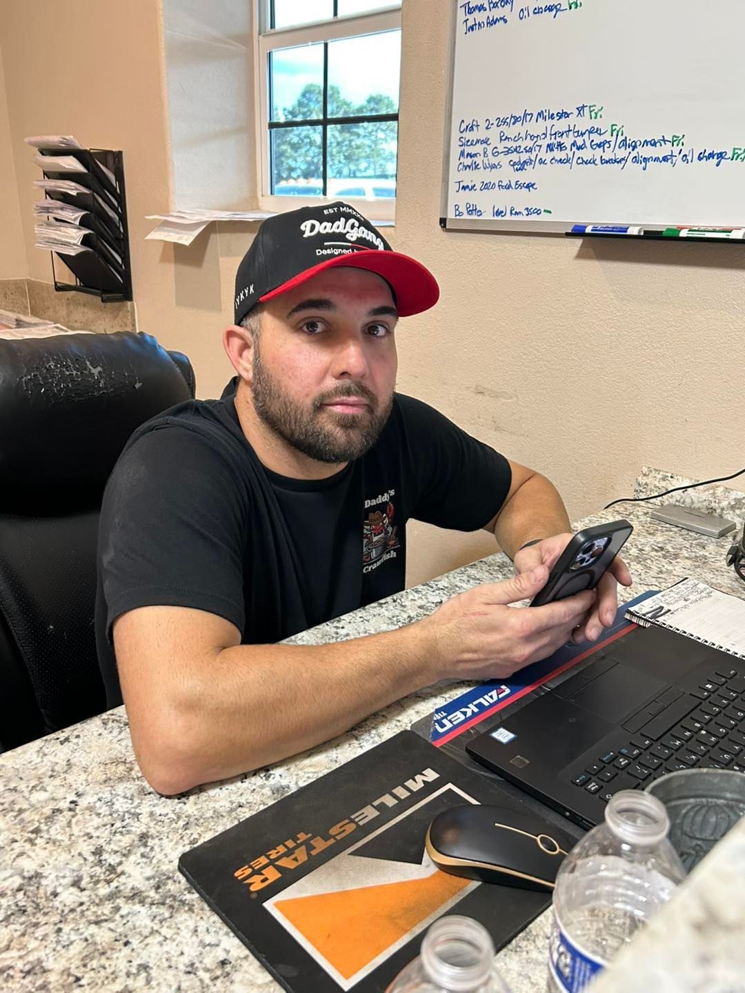 Man wearing a black hat and shirt, holding a phone, sitting at a desk with a laptop and mouse.