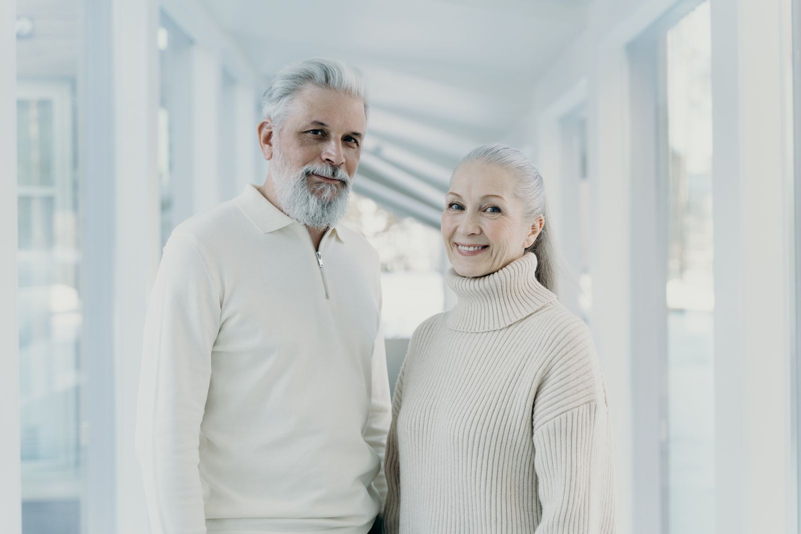 Two smiling people standing in a bright hallway, wearing light-colored sweaters.