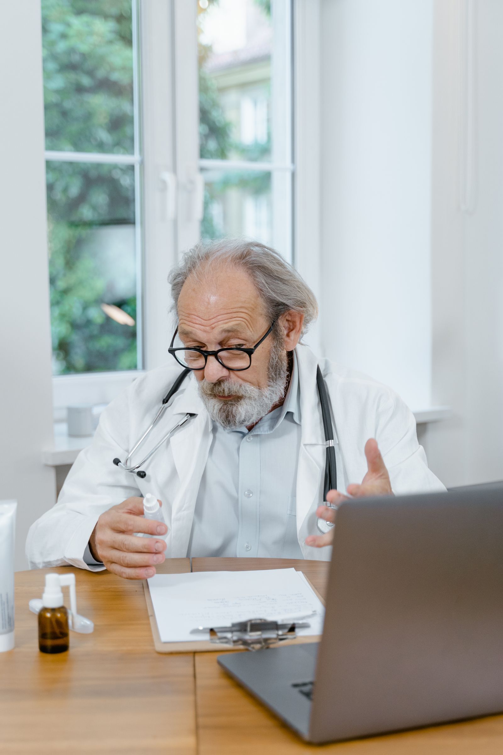 Doctor talking on a video call at a desk with a laptop and notes in a bright office