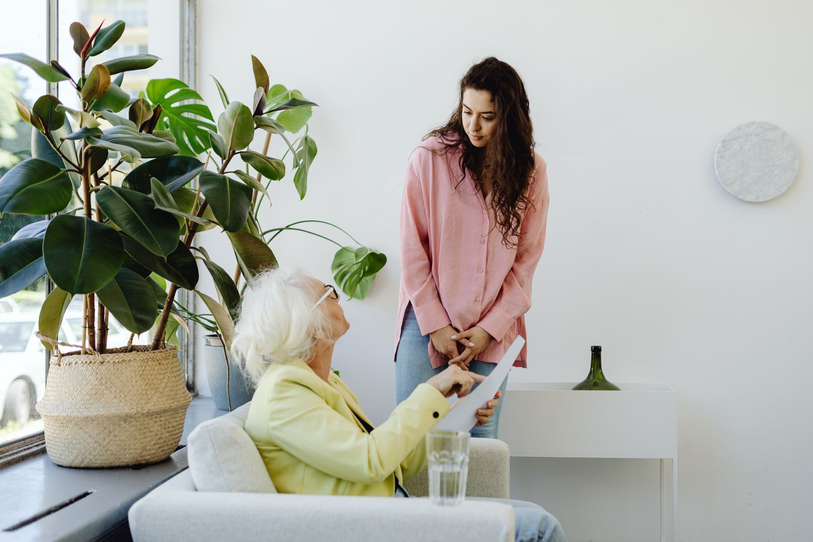 Two women talking in a bright living room, one seated on a sofa and one standing by a table with plants.
