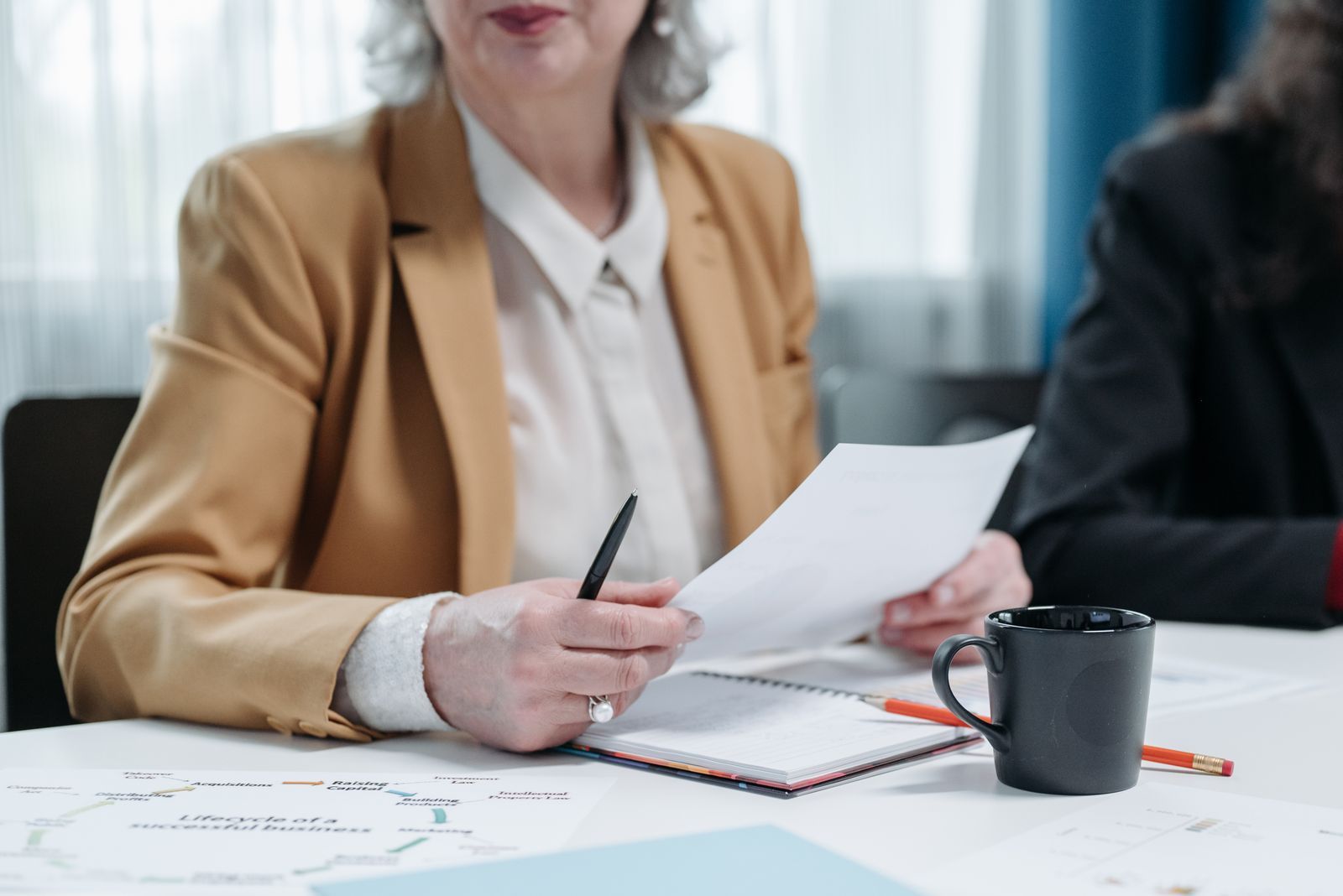 Business meeting with a person in a tan blazer reviewing papers at a table with a mug and notepad