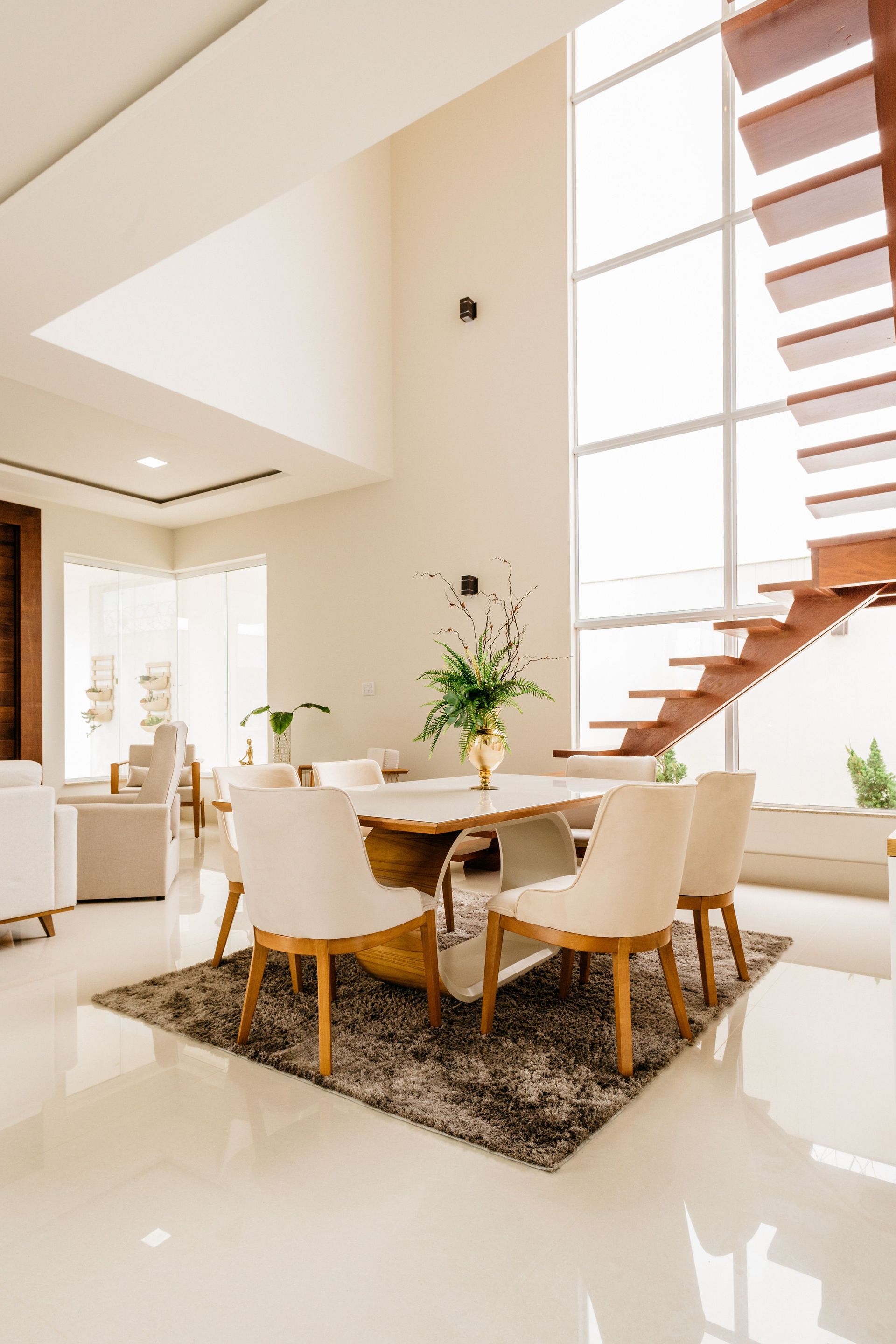 Bright modern dining area with a wooden table, white chairs, rug, and staircase by tall windows