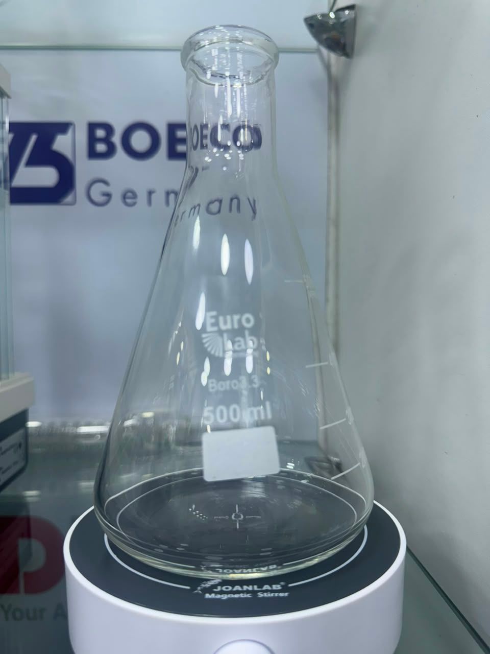 Matraz Erlenmeyer de vidrio transparente sobre un soporte de exhibición negro con un letrero de BOECO al fondo.