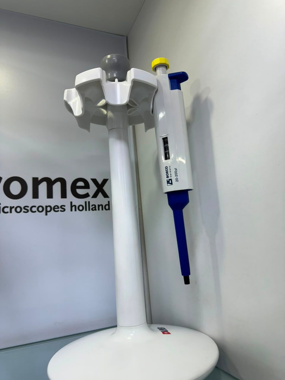 Soporte para microscopio de laboratorio blanco y azul sobre una base redonda, apoyado contra una pared gris 