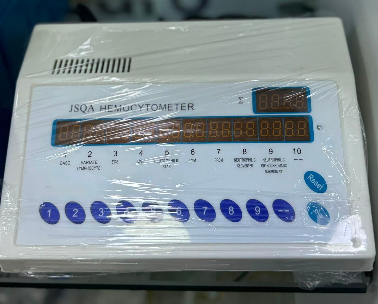 Dispositivo de conteo JSQ4 envuelto en plástico con botones azules y pantalla digital sobre una superficie blanca.