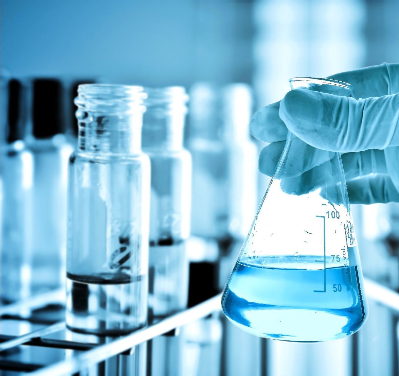 Mano enguantada sosteniendo un matraz con líquido azul en un laboratorio con tubos de ensayo de vidrio.