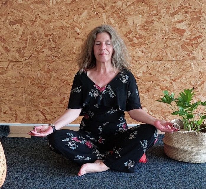 Vrouw in een jumpsuit met bloemenprint mediteert, zittend op een mat, ogen gesloten, handen open, plant in de buurt.