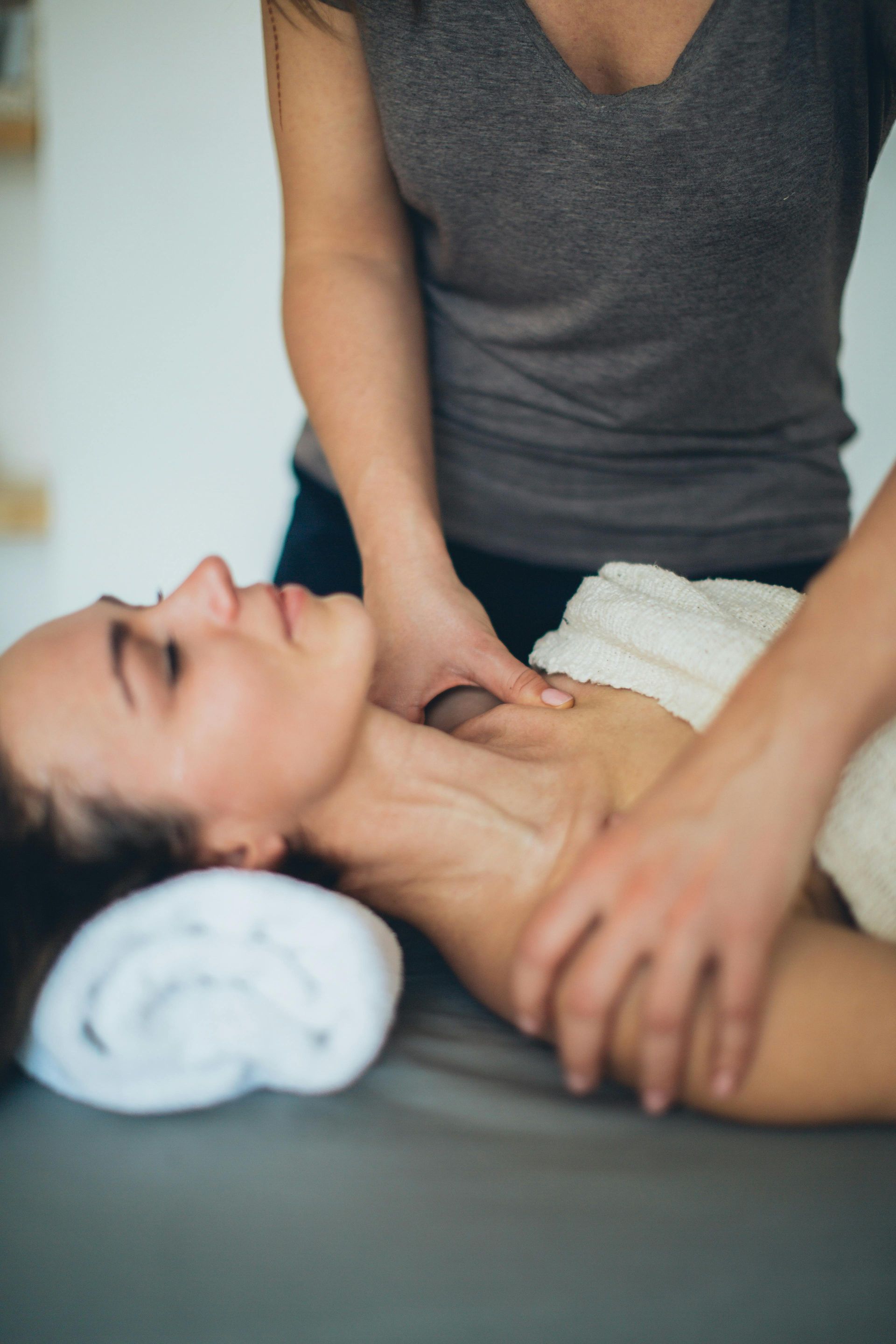 Een vrouw ligt op haar rug met gesloten ogen en krijgt een massage, terwijl de therapeut haar nek en schouder masseert.