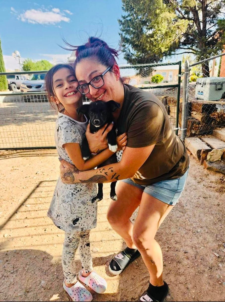 Woman and child hugging a small black puppy outdoors. They are smiling.