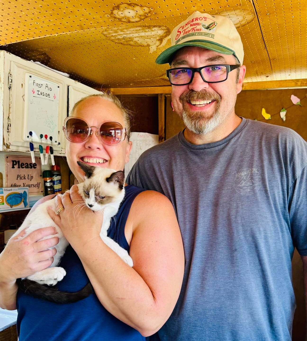 Woman holding a small white and tan cat smiles with a man in a shed.