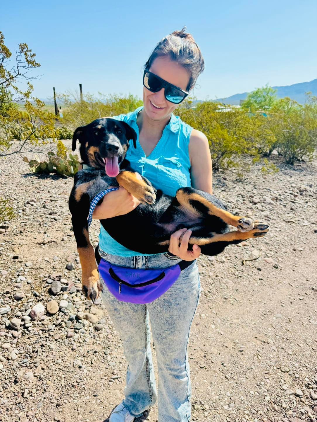 Woman holding a smiling black and brown dog outdoors in a desert setting; sunny day.