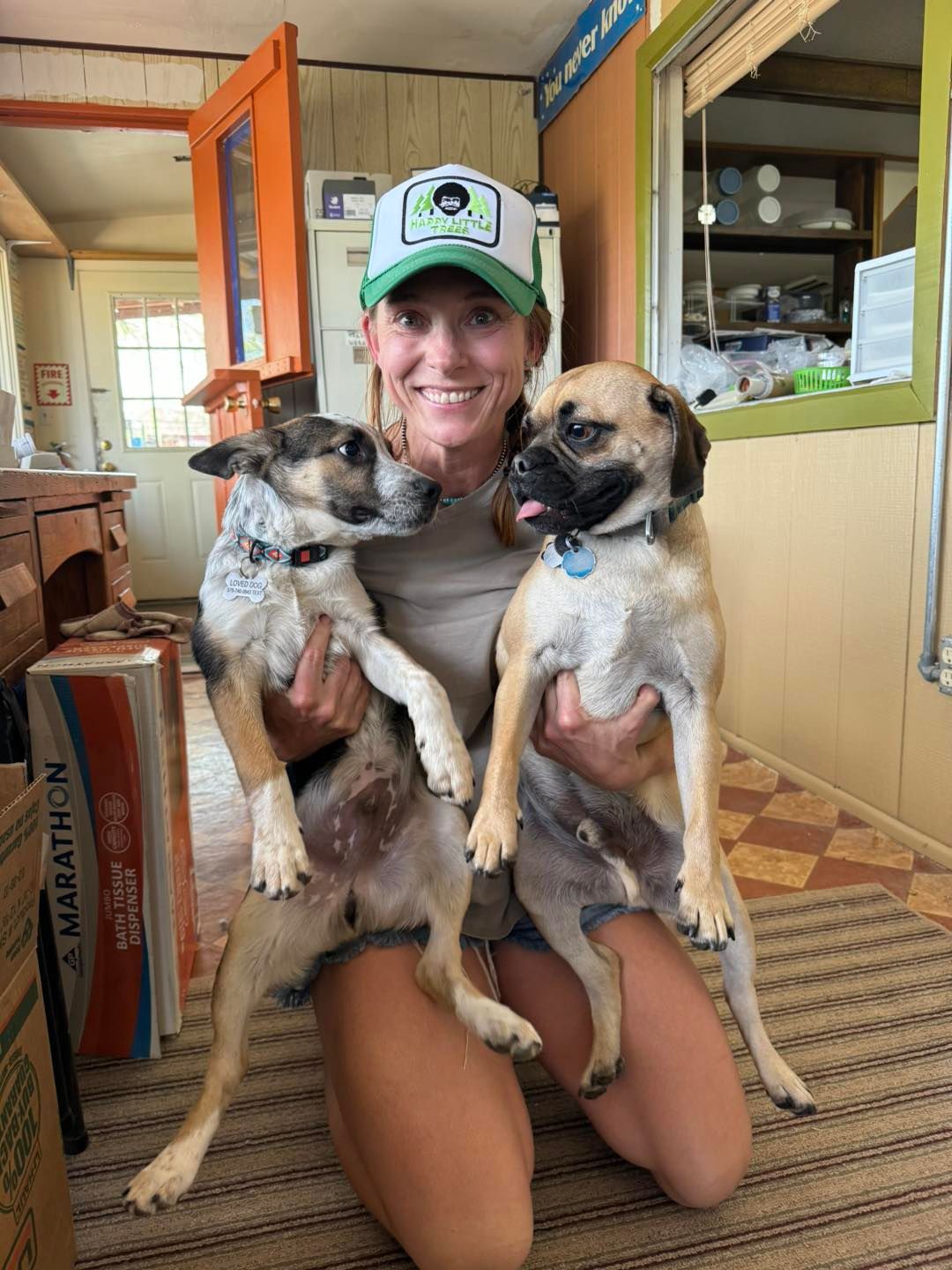 Woman kneeling, holding two dogs. Interior setting, smiling, dogs looking at camera.