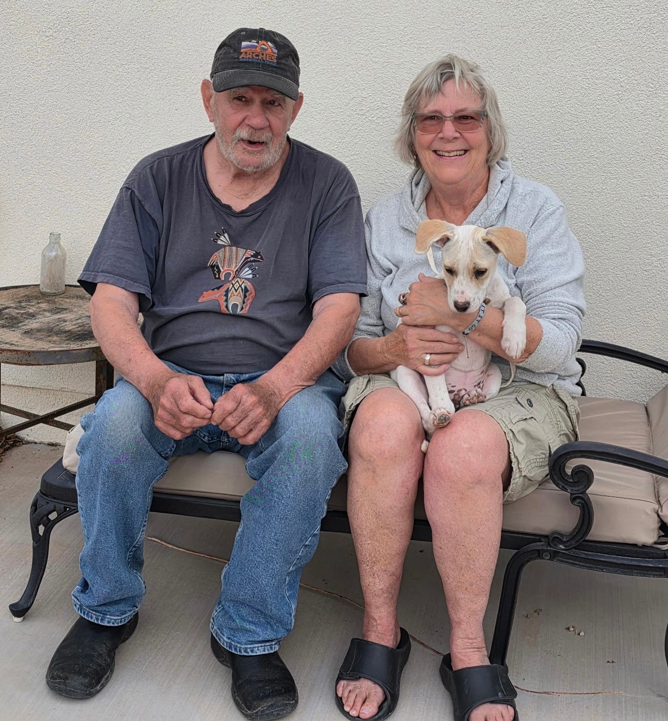 Two people sit on a bench holding a small white-and-tan dog against a plain wall.