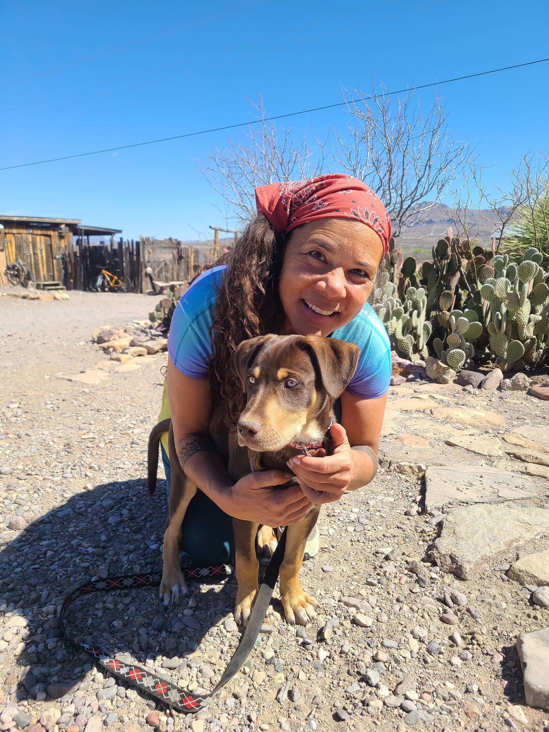 A person with long, curly hair and a red headscarf kneels in a desert setting, smiling while hugging a brown dog on a leash.