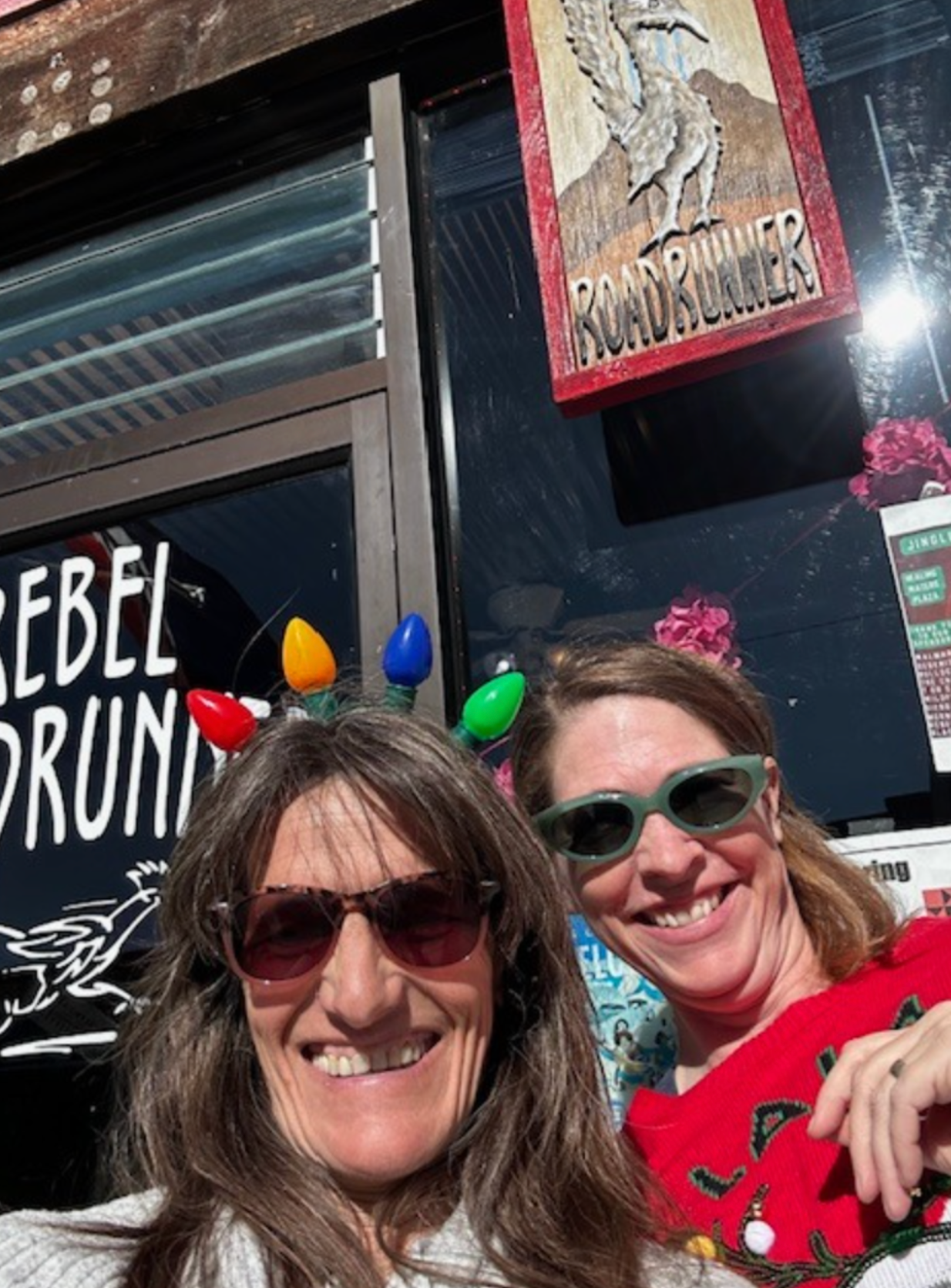 Two smiling people wearing sunglasses and festive lights in front of a bar sign.