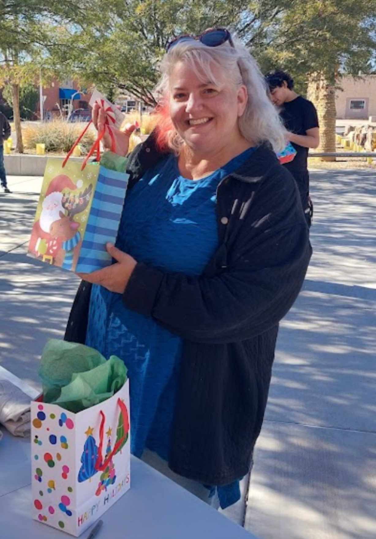 Woman smiling, holding gift bags outdoors, blue shirt and black jacket.